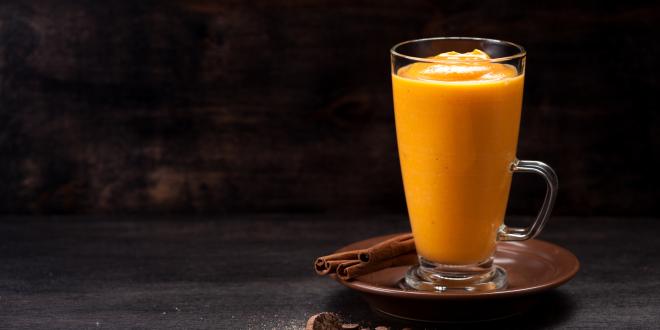 A glass cup filled with pumpkin spice smoothie next to cinnamon sticks. 